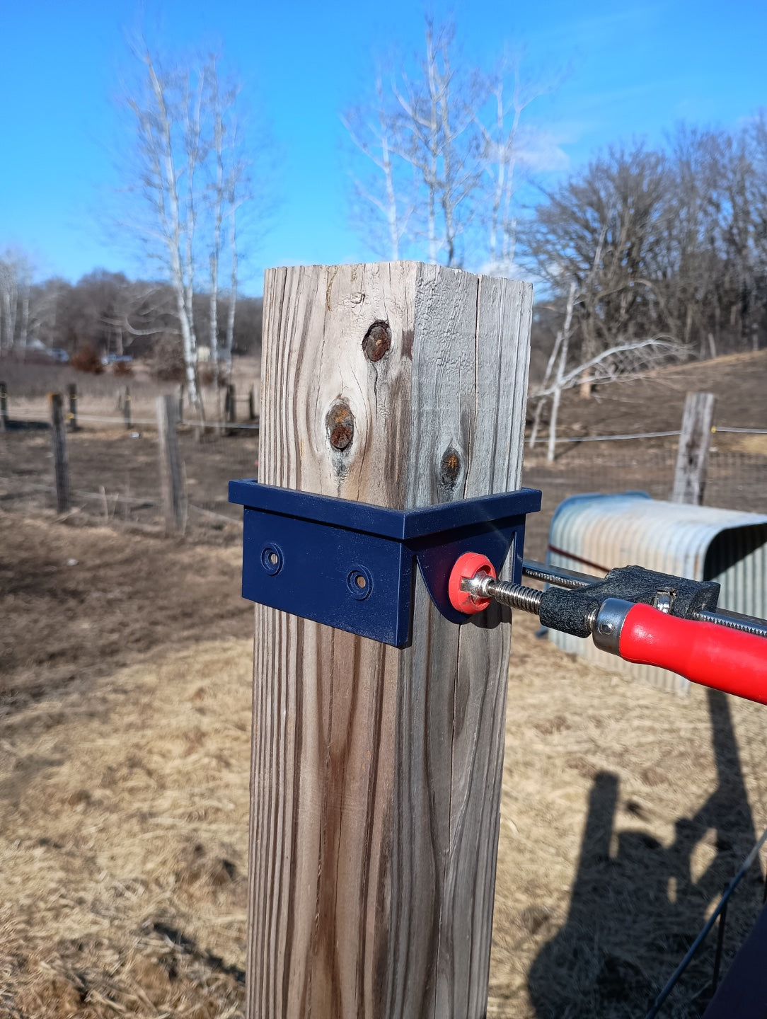Black 3D printed 4x4 circular saw cutting guide clamped around a wooden fence post, viewed from the opposite side with red clamp handle