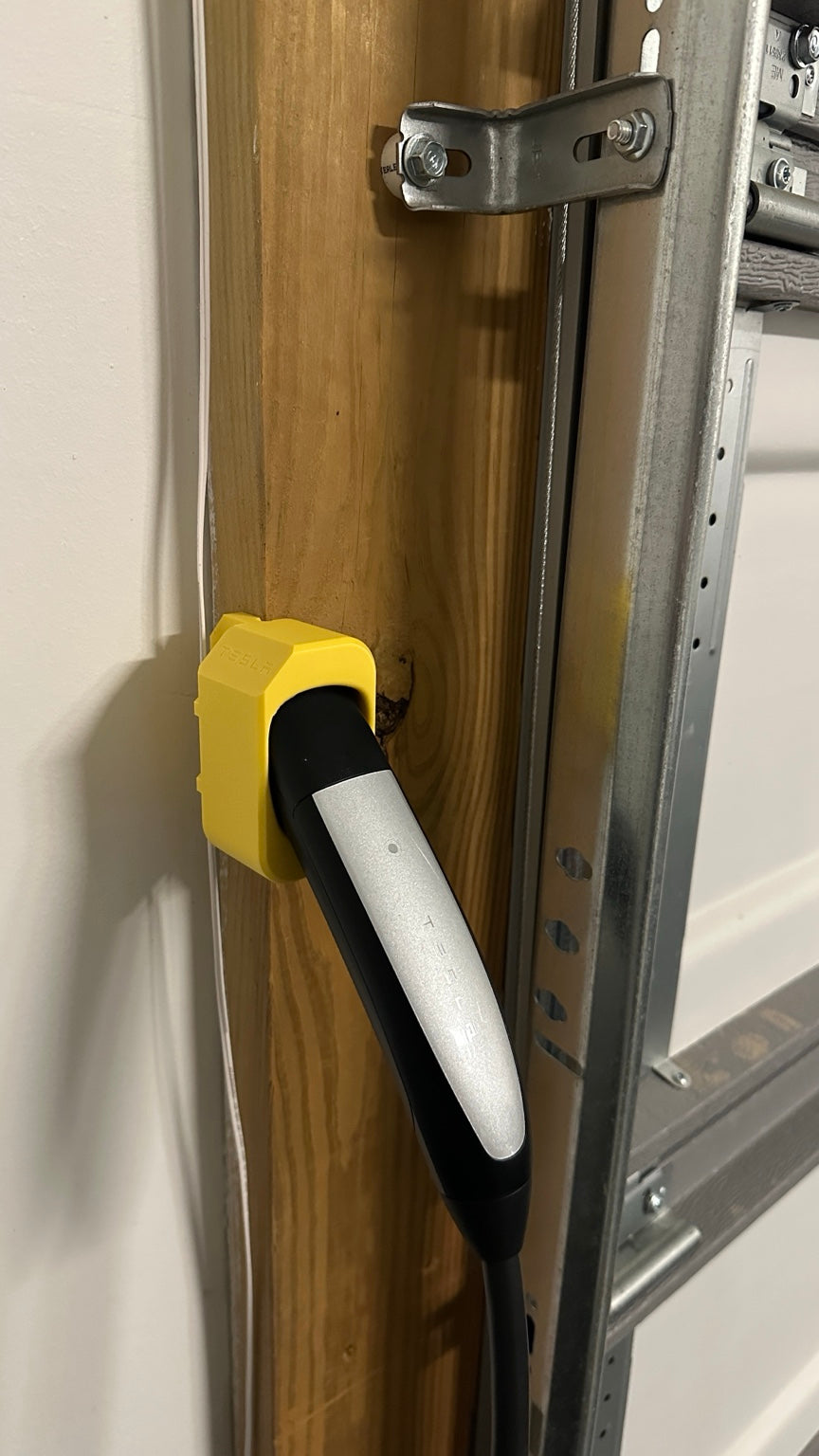 Yellow prototype NACS charging dock installed on wooden garage corner with Tesla charging cable parked in holder